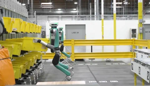 The previous generation of Agility's Digit robot, featuring its iconic backward-bending legs, handling totes in a warehouse.